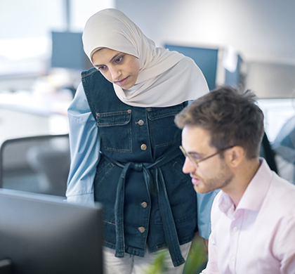 In Worley office, woman standing next to a young man at a desk both looking at the computer.
